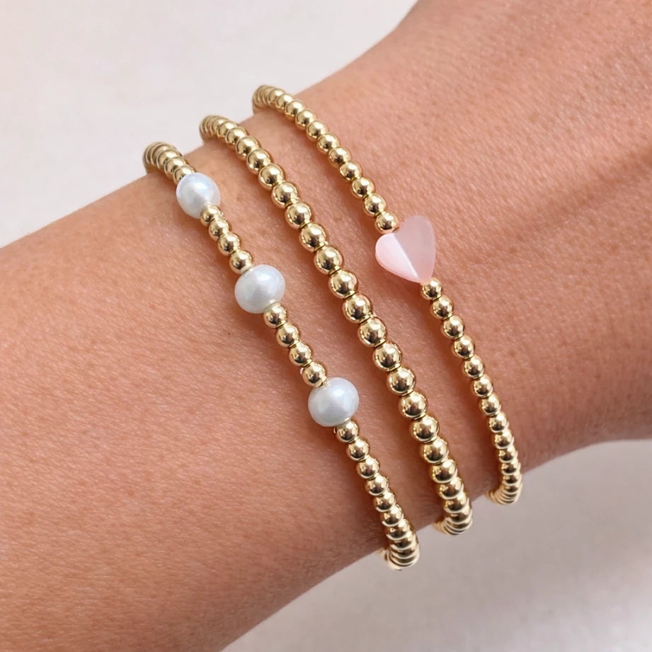Gold beaded bracelets with white and pink heart beads on a wrist against a light background