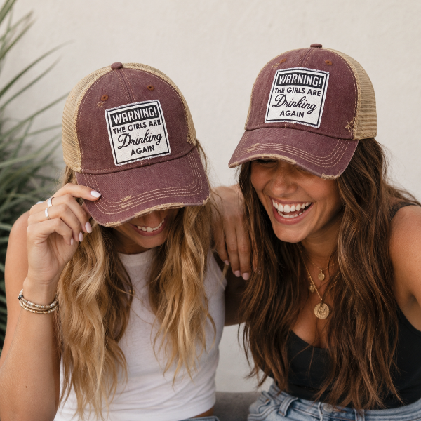 Two women wearing matching distressed trucker hats with a maroon front panel, tan mesh back, and a white patch that reads 'WARNING! THE GIRLS ARE Drinking AGAIN'.