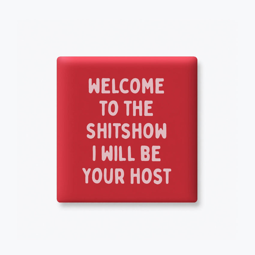 A square red pinback button or badge with rounded corners, featuring the text 'WELCOME TO THE SHITSHOW I WILL BE YOUR HOST' in white hand-drawn style lettering, shown on a light gray background.