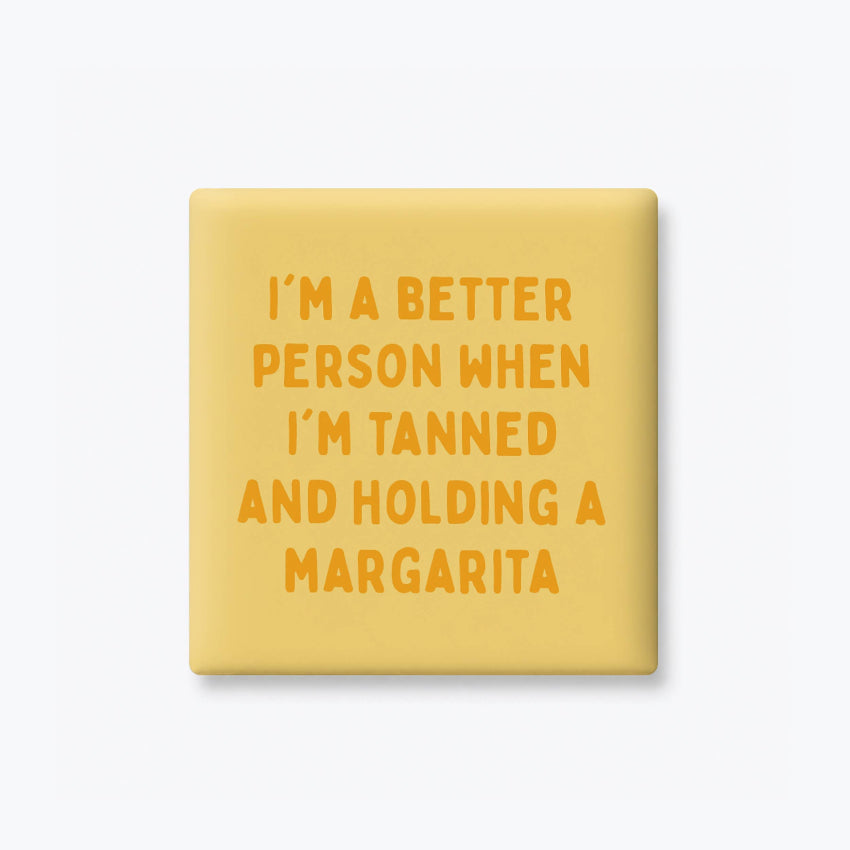 A square yellow pinback button or badge with orange text that reads, 'I'M A BETTER PERSON WHEN I'M TANNED AND HOLDING A MARGARITA', shown on a light gray background.